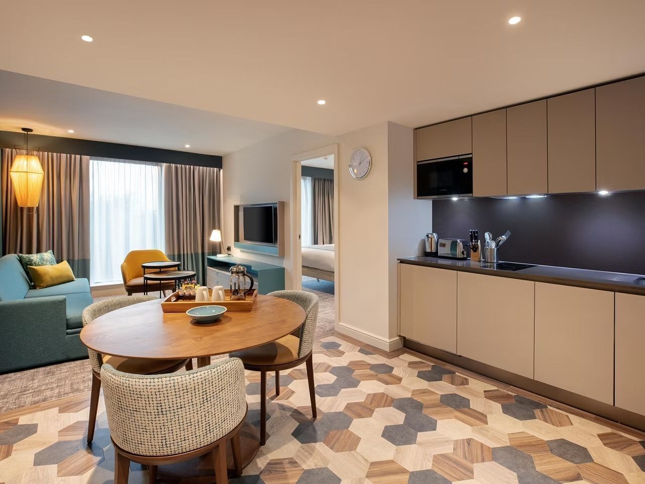 A modern hotel room with a dining area, a sofa, a TV, and a kitchen area.