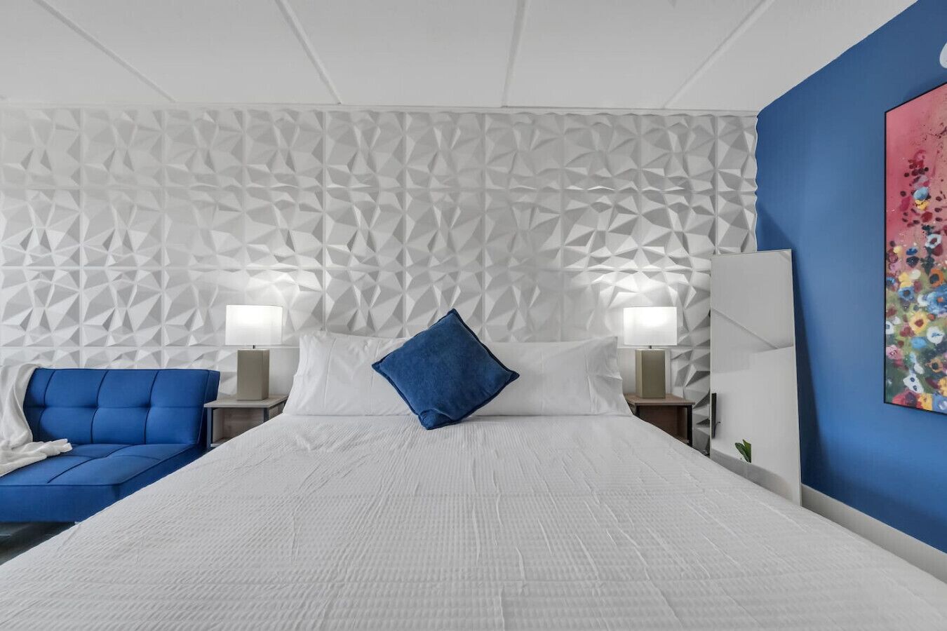 Ein modernes Hotelzimmer mit einem Kopfteil im geometrischen Muster, einem großen Bett mit weißen Bettwäsche, einem blau gepolsterten Sofa, zwei Nachttischen mit Lampen und einem farbenfrohen abstrakten Gemälde an der Wand.