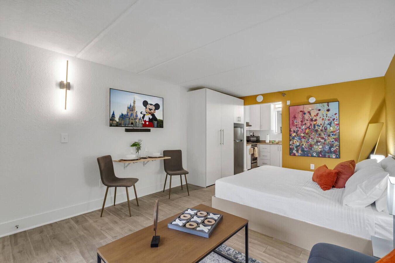 Moderni hotellihuone, jossa on sänky, työpöytä kahdella tuolilla, pieni keittokomero ja suuri ikkuna.