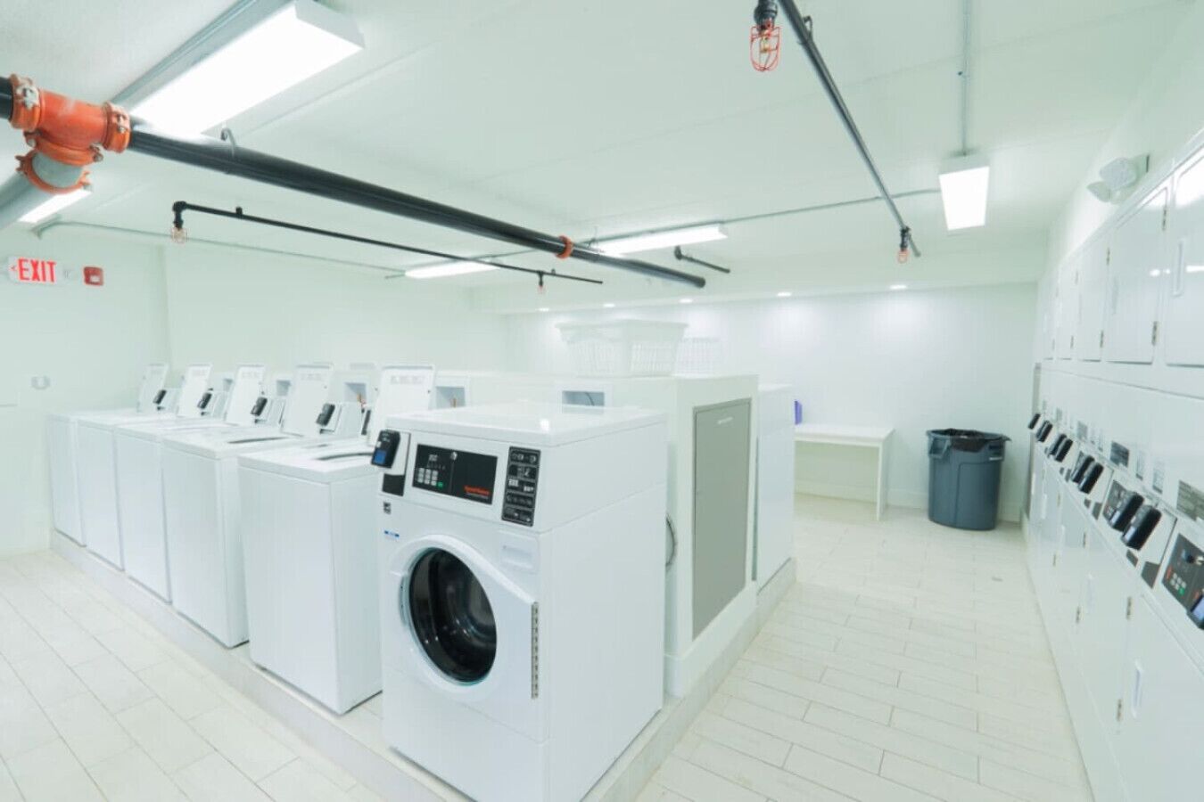 Une buanderie équipée de plusieurs machines à laver et de plusieurs sèche-linge, avec un panneau indiquant la sortie et une poubelle.
