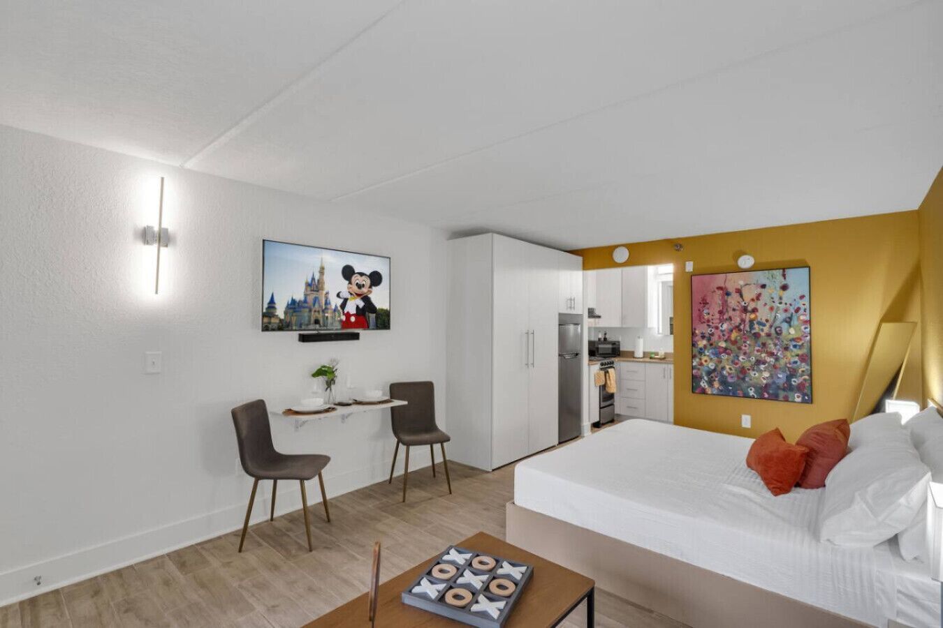 A hotel room with a bed, a desk with chairs, a television, a kitchen area, and a colorful painting.