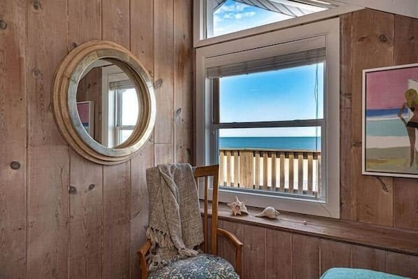 Ein Zimmer mit Holzboden und einem großen Fenster mit Meerblick, ein runder Spiegel und ein Sessel mit Decke.