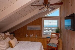 Ein gemütliches Schlafzimmer im Dachgeschoss mit schrägen Wänden, einem Deckenventilator, einem großen Fenster mit Jalousien, einem Bett mit Kissen, einem Sessel und einem an der Wand befestigten Fernseher.