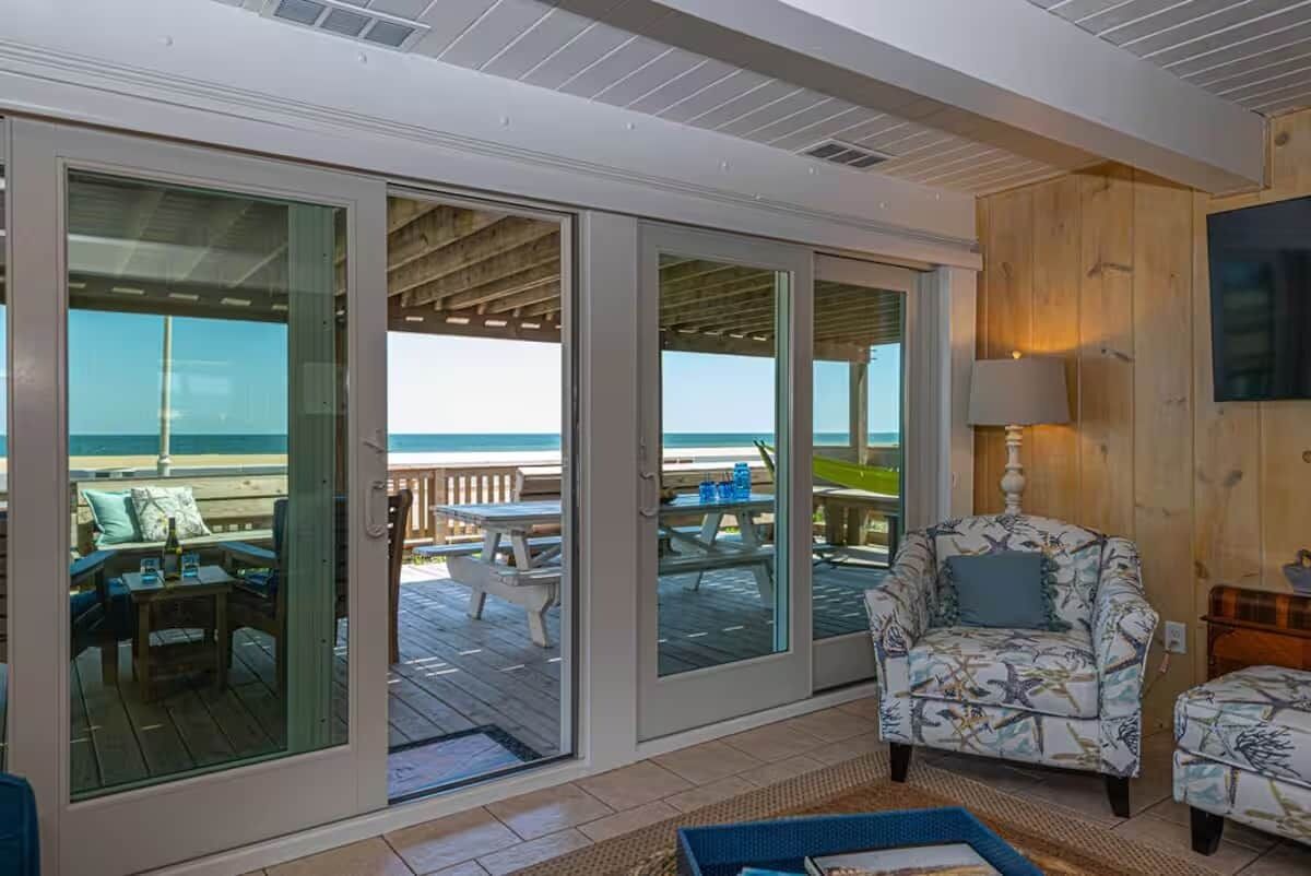 A room with a view of the ocean, a wooden deck, and a seating area with a patterned armchair and ottoman.