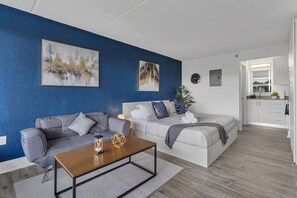 Uma sala de estar moderna com um sofá, uma mesa de centro e uma cama com cabeceira. Há duas pinturas na parede, um relógio e uma planta.