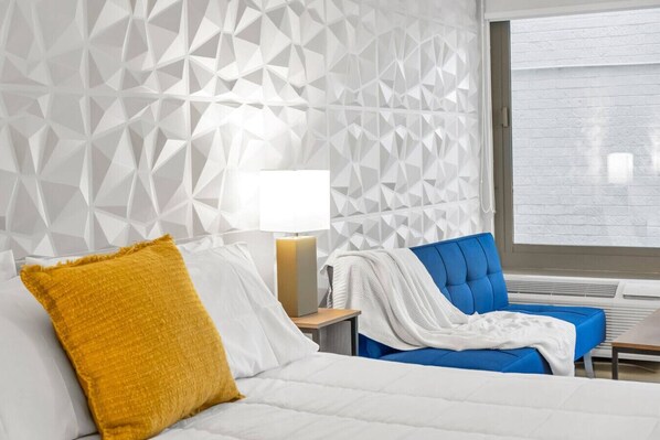 Une chambre d’hôtel avec un mur orné d’un motif géométrique, un lit avec une literie blanche et un plaid de couleur moutarde, un fauteuil bleu recouvert de tissu, une table de chevet avec une lampe et une fenêtre avec des stores.