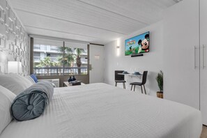 A modern hotel room with a large bed, a desk with two chairs, a television on the wall, and a window with a view of palm trees. - Charming Apt 7 min close to Disney + Free Parking (Kissimmee)