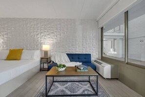 Een moderne hotelkamer met een witte, geribbelde wand, een blauwe bank, een houten salontafel met een plant en boeken, een witte nachtkastje met een lamp, en een groot raam met uitzicht op het interieur van het hotel.