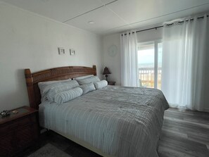 A bedroom with a wooden headboard, a bed with grey bedding, a nightstand, a lamp, and a view of the ocean through a window with white curtains. - Beachfront House - Sunny Beach Retreat (Gilchrist)