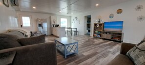 A modern living room with a sofa, coffee table, and TV. There is a kitchen area with a fridge and microwave. The room has wooden flooring and a view of the outdoors. - Beachfront House - Sunny Beach Retreat (Gilchrist)