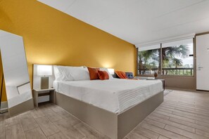 Ein Hotelzimmer mit einem großen Bett, einem Nachttisch, einem Fenster mit Blick auf Palmen und einer Tür.