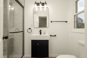 Un baño con cabina de ducha de vidrio, un lavamanos blanco con mueble negro, un espejo con iluminación y una ventana.