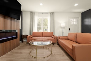 A modern living room with a sofa set, a glass coffee table, and a television mounted on a wooden panel. - Rockne Retreat<>6BR 3BA Home Close to ND (South Bend)