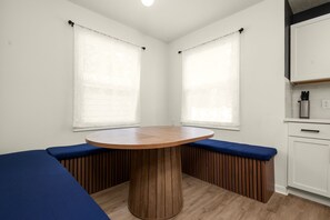 Una habitación con una mesa redonda de madera rodeada de bancos acolchados azules, paredes blancas y una zona de cocina con armarios blancos.