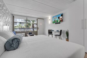 A modern hotel room with a large bed, a desk with two chairs, a television mounted on the wall, and a window with a view of palm trees. - Family Suite 7 Min from Disney w Free Parking (Kissimmee)