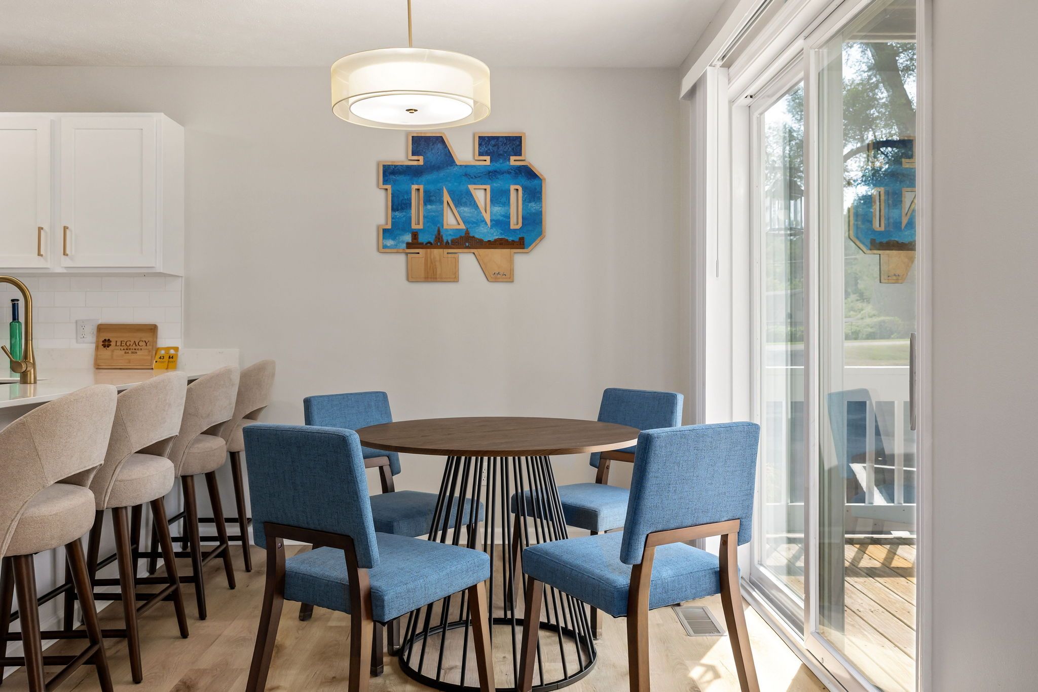 A dining area with a round table surrounded by blue chairs, white cabinets, and a large window.