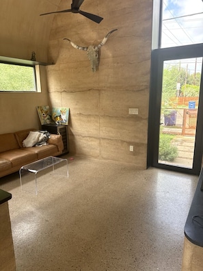 Ein Zimmer mit Betonboden, Deckenventilator, großem Fenster, braunem Ledersofa und einer Wandskulptur.