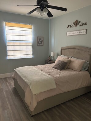 Ein Schlafzimmer mit Bett, Deckenventilator, Fenster mit Jalousien, Nachttisch mit Lampe und Wanddekoration.