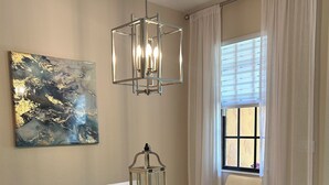 A dining area with a glass-top table, white chairs, a wooden bench, a hanging light fixture, a wall-mounted abstract painting, and a window with curtains. - OLE’ Lely resort. Beautiful 3-bedroom 2.5 bath townhome. Resort-style living! (Naples)