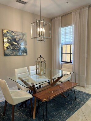 Un comedor con mesa de vidrio, sillas blancas, un banco de madera, una lámpara colgante, un cuadro abstracto en la pared y una ventana con cortinas.