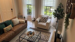 A living room with a brown leather sofa, a white armchair, a glass coffee table with a board game, and a view of greenery through the windows. - OLE’ Lely resort. Beautiful 3-bedroom 2.5 bath townhome. Resort-style living! (Naples)