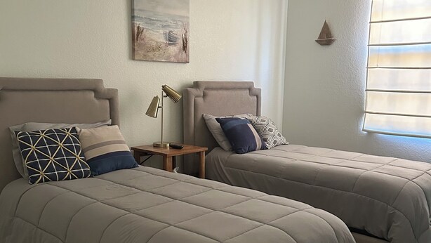 A bedroom with two beds, a nightstand, a lamp, a painting, and a window with blinds. - OLE’ Lely resort. Beautiful 3-bedroom 2.5 bath townhome. Resort-style living! (Naples)