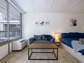 Ein modernes Wohnzimmer mit einer Couch, einem Couchtisch und einem Bett. Drei Wandbilder und ein Fenster mit Blick auf ein Gebäude sind ebenfalls vorhanden.