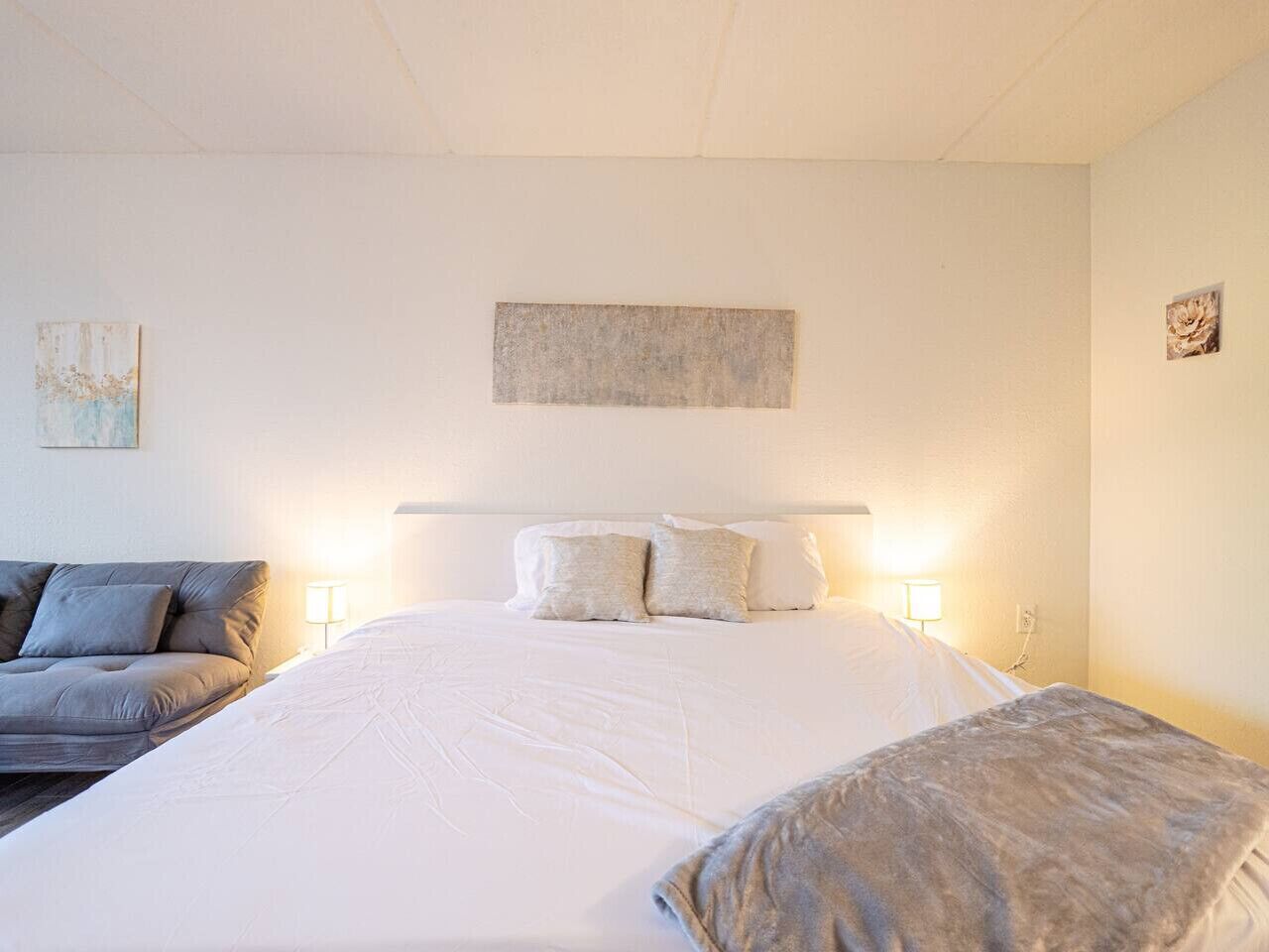 Ein Schlafzimmer mit einem großen Bett, zwei Nachttischlampen, einem grauen Sofa und einem kleinen Gemälde an der Wand.