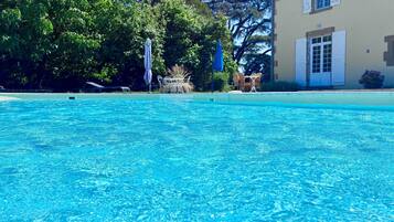 Une piscine d’un bleu éclatant, dotée d’un parasol et de chaises longues, entourée d’arbres et bordée d’un bâtiment à la façade beige.