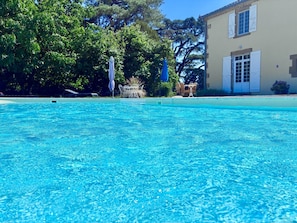 En krystalklar pool med parasol og liggestole, omgivet af træer og en bygning med beige facade.