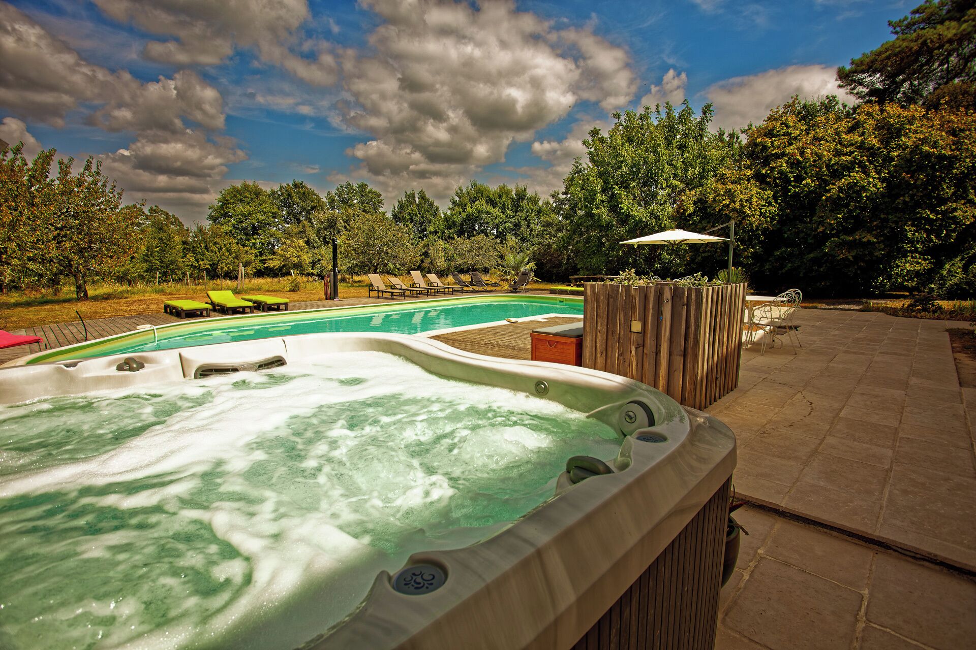 Un bain à remous doté d’une terrasse en bois et de chaises longues, entouré d’arbres et bordé d’une piscine.