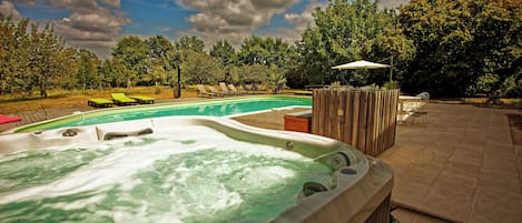 Un jacuzzi con terraza de madera y sillas reclinables, rodeado de árboles y una piscina.