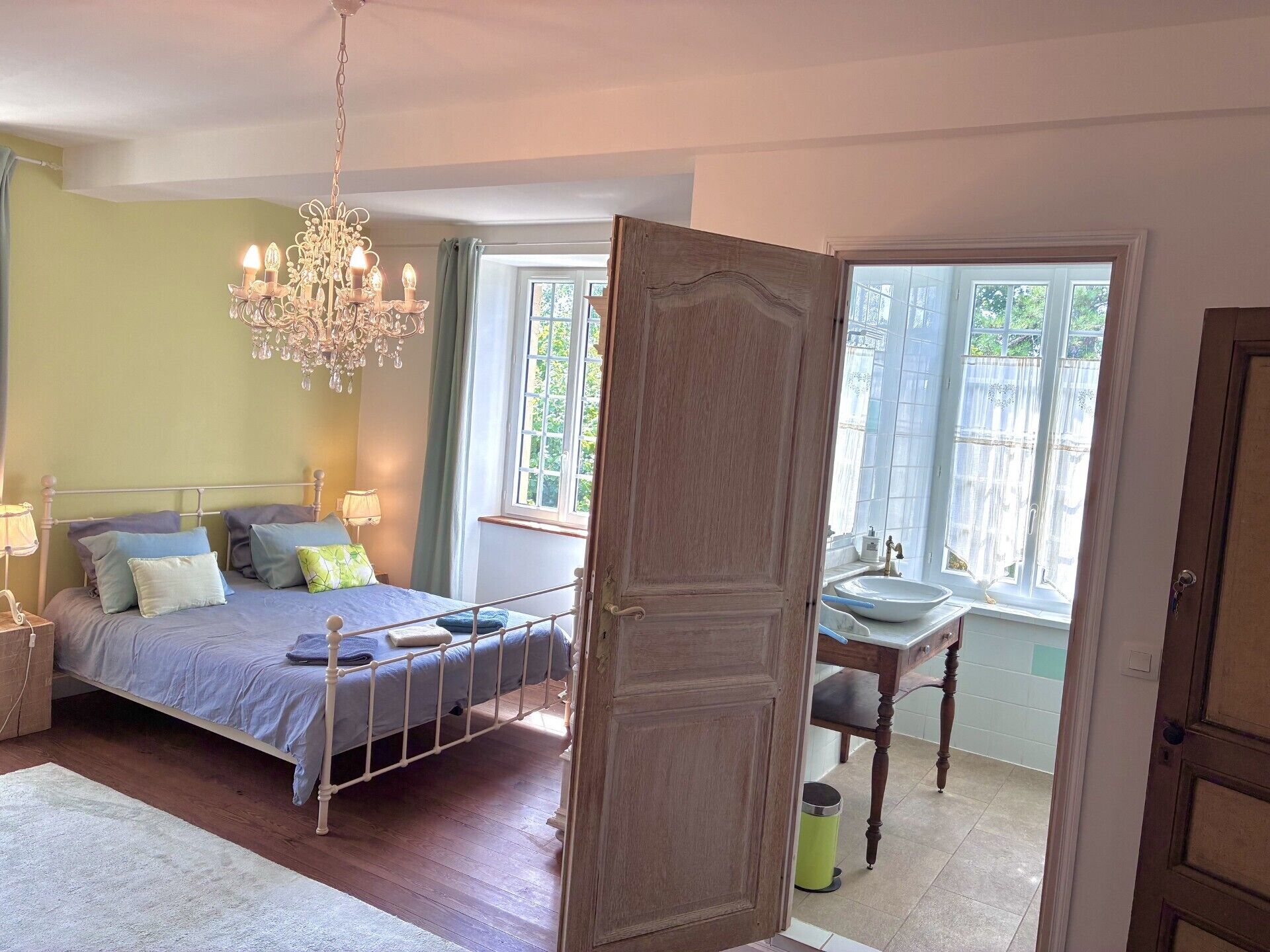 A bedroom with a bed, bedside tables, a chandelier, and a view of the outdoors through a window.