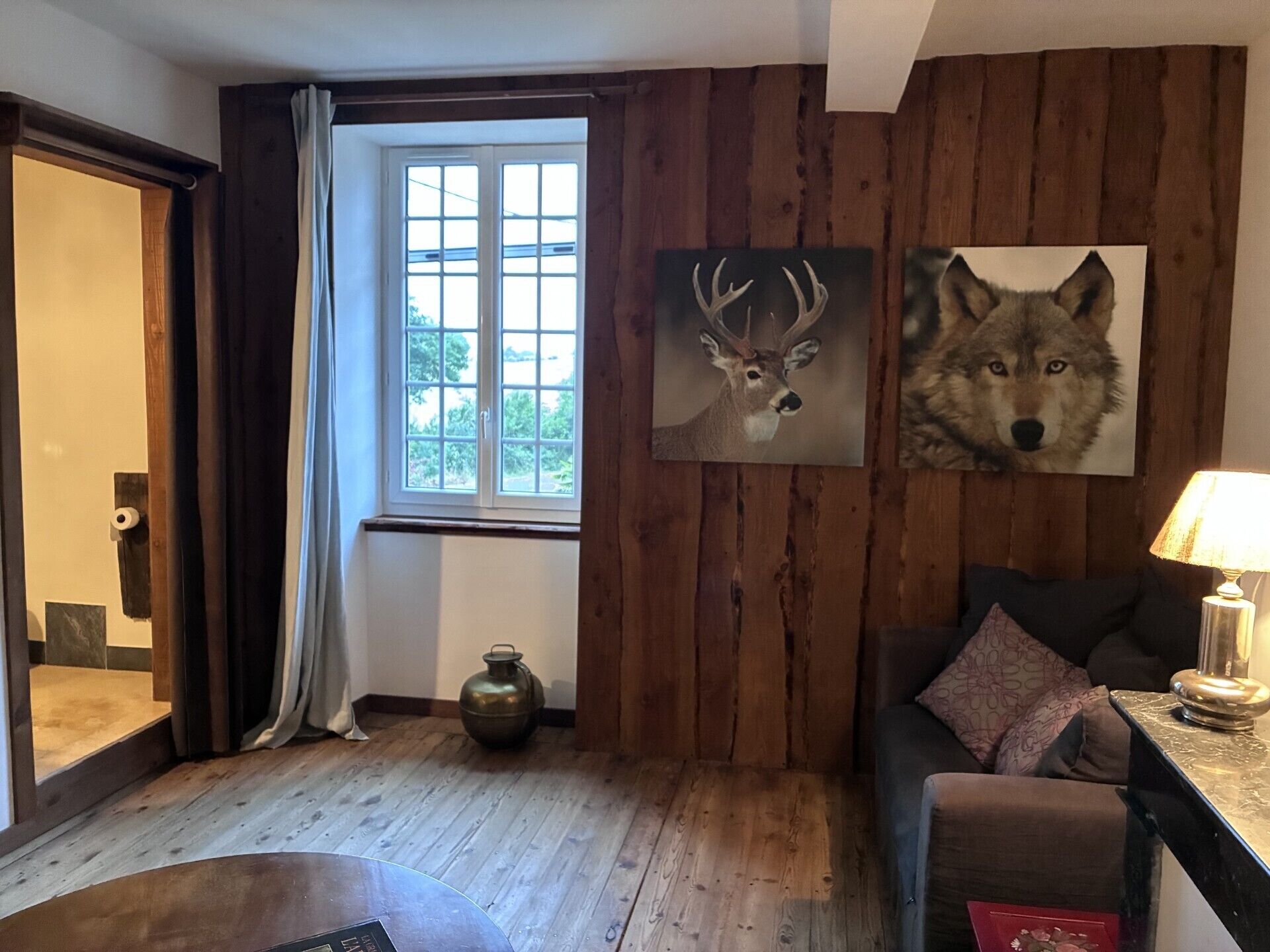 A cozy room with wooden walls, a window with curtains, a sofa with cushions, a table with a lamp, and two wall-mounted animal paintings.