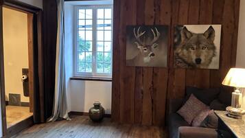 A cozy room with wooden walls, a window with curtains, a sofa with cushions, a table with a lamp, and two wall-mounted animal paintings.