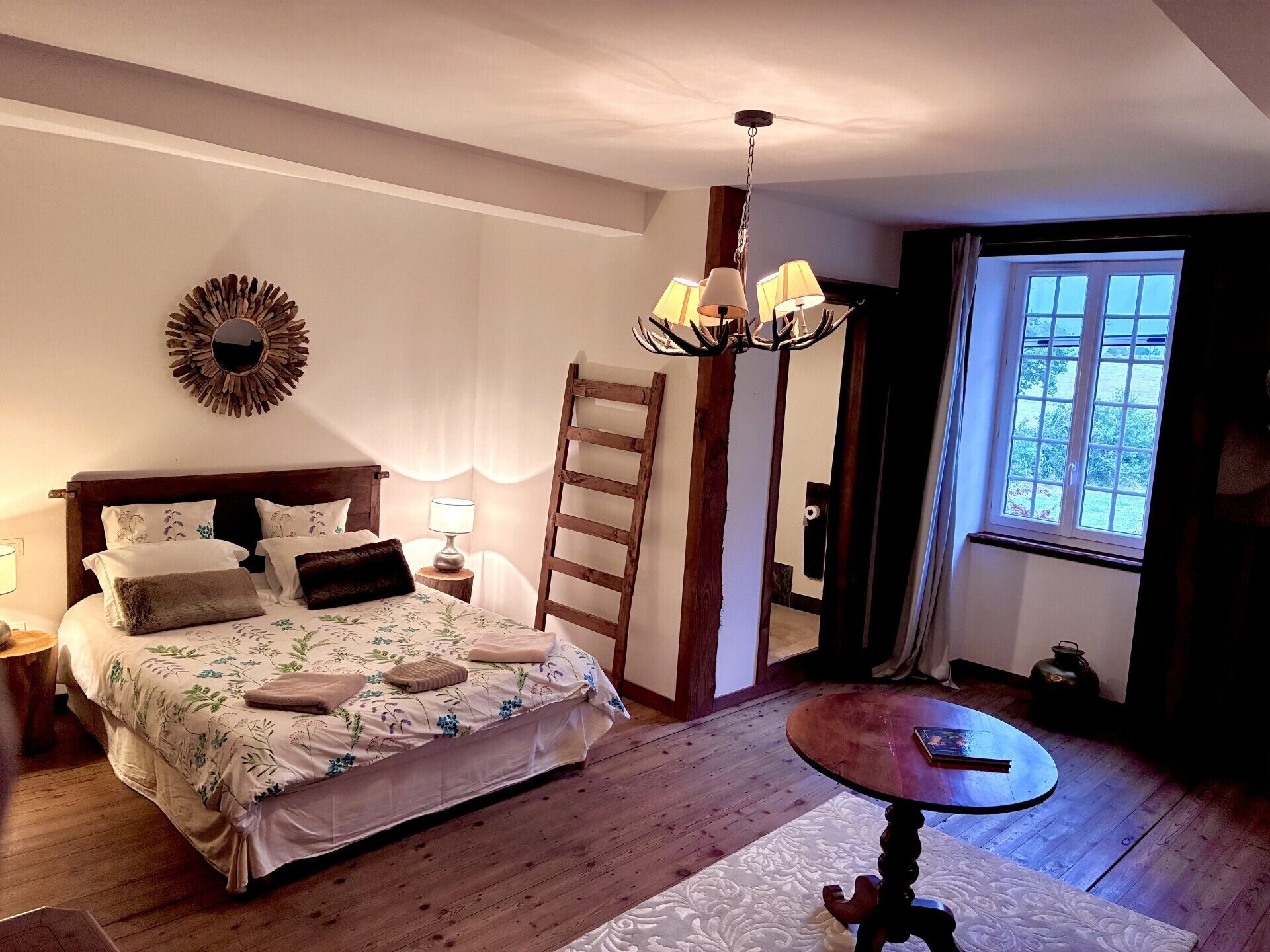 Une chambre à coucher avec un lit, des lampes de chevet, une chaise en bois, une table ronde et une fenêtre avec des rideaux.