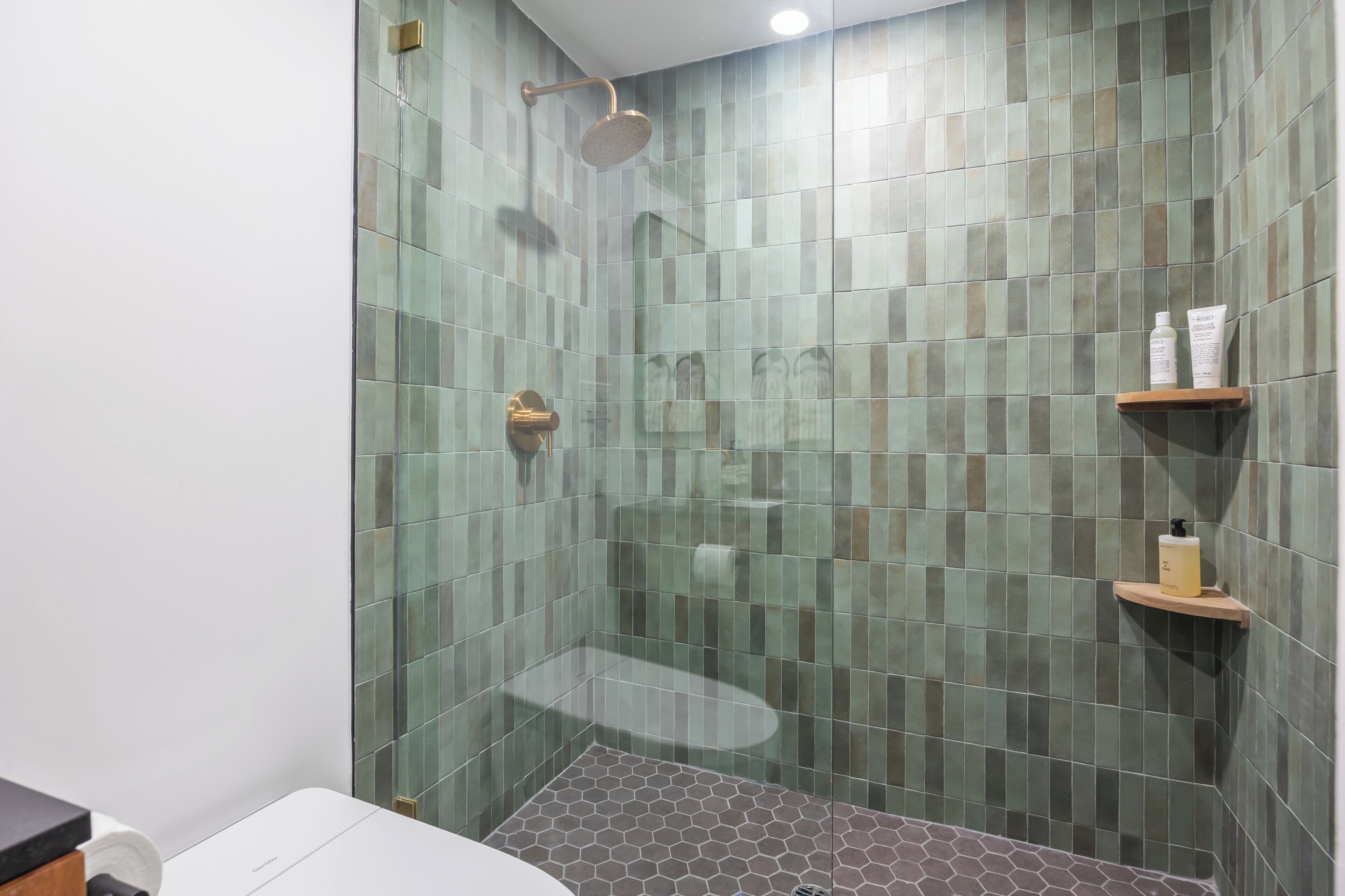A shower area with a glass door, a handheld showerhead, and a shelf with toiletries.