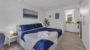 Ett sovrum med en säng, ett nattduksbord, en lampa och utsikt över köket.