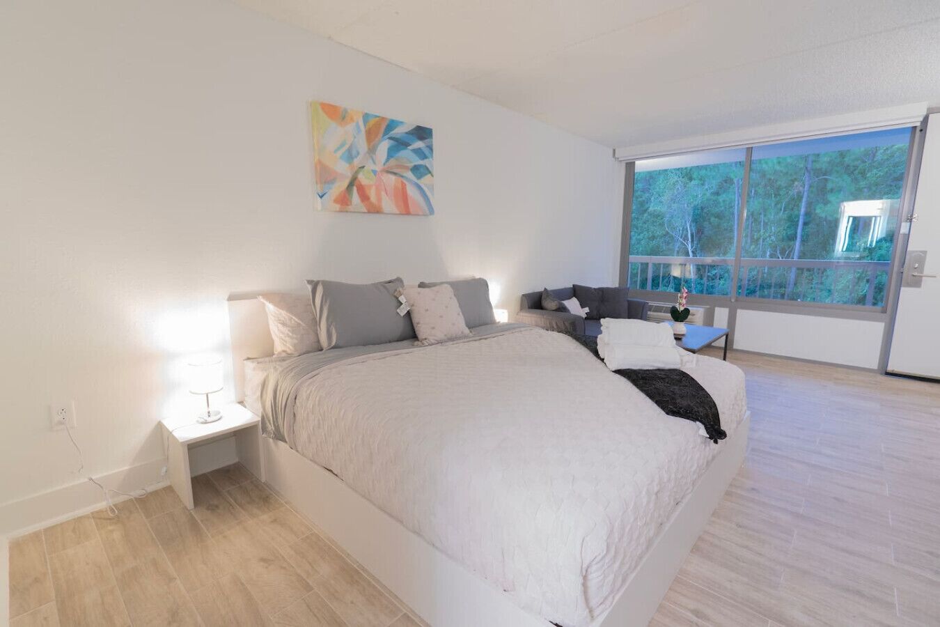 A bedroom with a large bed, a nightstand, a lamp, a window with a view of trees, and a painting on the wall.