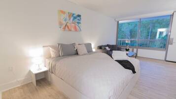 A bedroom with a large bed, a nightstand, a lamp, a window with a view of trees, and a painting on the wall.