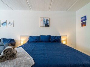 Une chambre d’hôtel avec un grand lit, un canapé, une lampe fixée au mur et une petite table sur laquelle est posée une serviette enroulée.