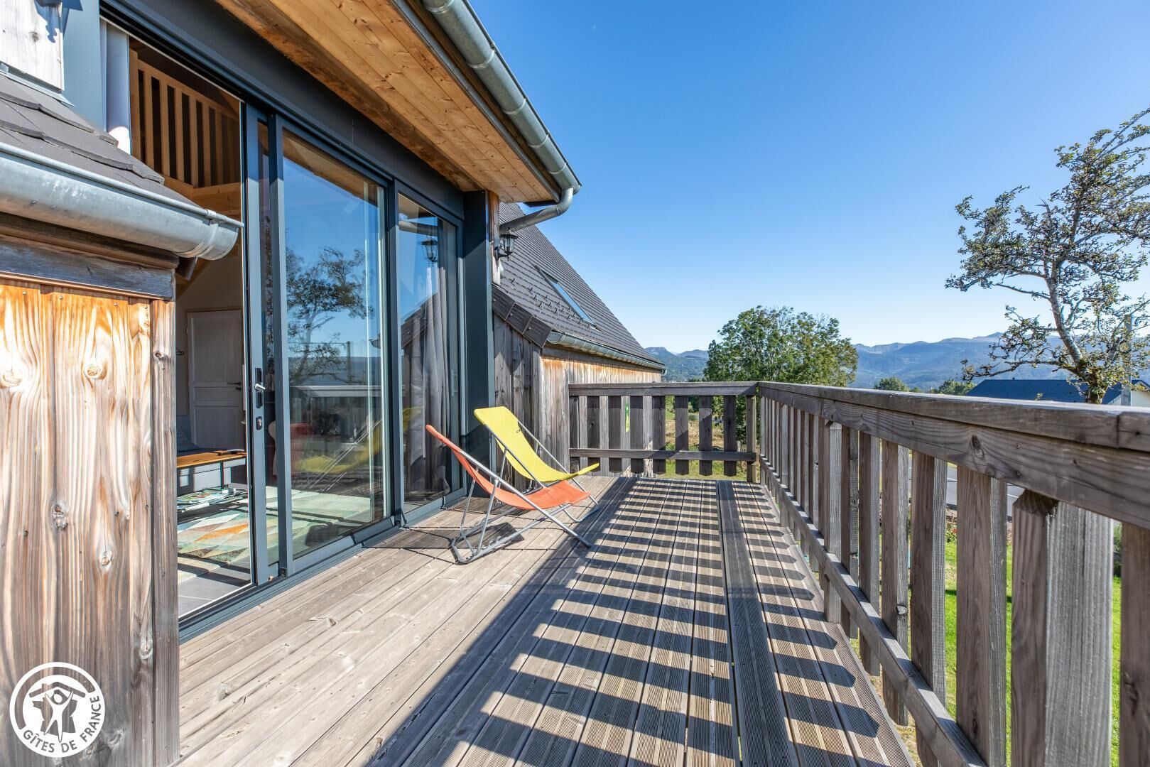 A wooden deck with a view of mountains, a glass door entrance, and two lounge chairs.