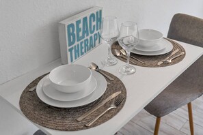 A dining table set with white plates, bowls, cutlery, and glasses, with a 'BEACH THERAPY' sign in the background. - Cozy Modern King Room Close to Disney + Pool | Orl (Kissimmee)