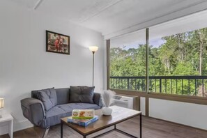 A room with a grey sofa, a table with a book, a floor lamp, and a large window with a view of trees. - Cozy Suite 7 Mins to Disney World + Free Parking (Kissimmee)