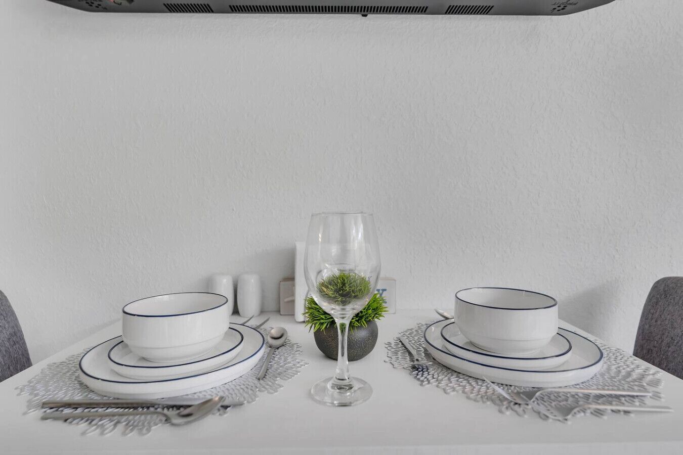 Une table dressée avec de l’assiette et des bols blancs, un verre d’eau, et une petite plante au centre.