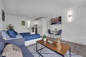 A modern living room with a sofa, a dining table, and a TV mounted on the wall. - Cozy Disney Escape at 8 min + Pool & Parking | Orl (Kissimmee)