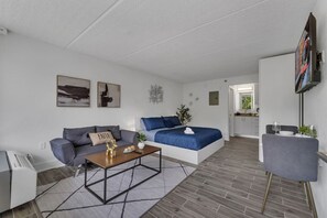 A modern living room with a sofa, a coffee table, and a bed. There is a television mounted on the wall and a kitchen area visible through an open door. - Cozy Disney Escape at 8 min + Pool & Parking | Orl (Kissimmee)