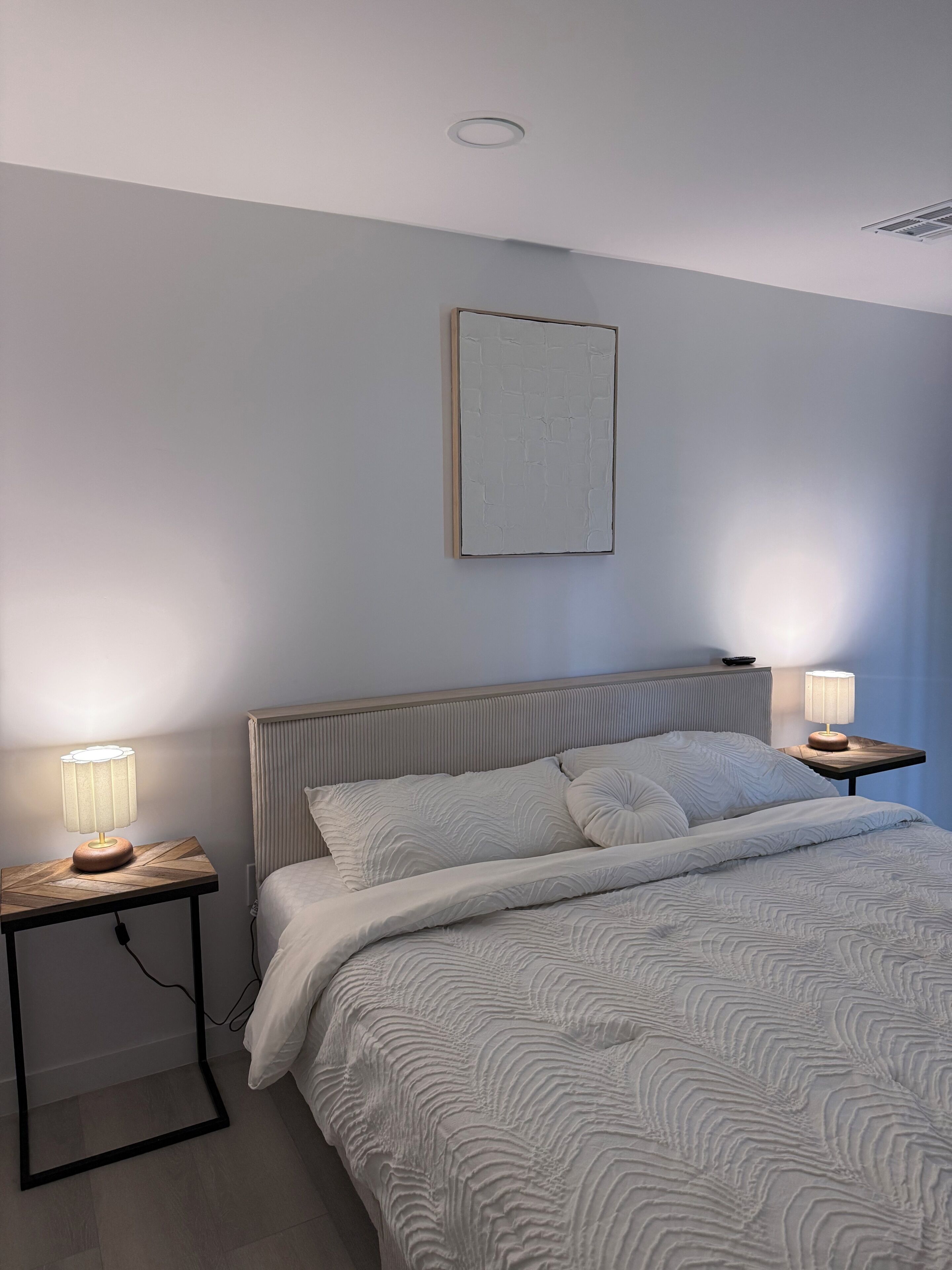 Un dormitorio con una cama, dos mesitas de noche con lámparas, un radiador y un cuadro enmarcado en la pared.