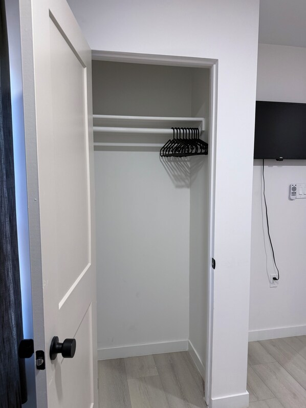 A room with a white door, a black door handle, a white closet with a black hanger rack, a flat-screen TV mounted on the wall, and a power outlet.