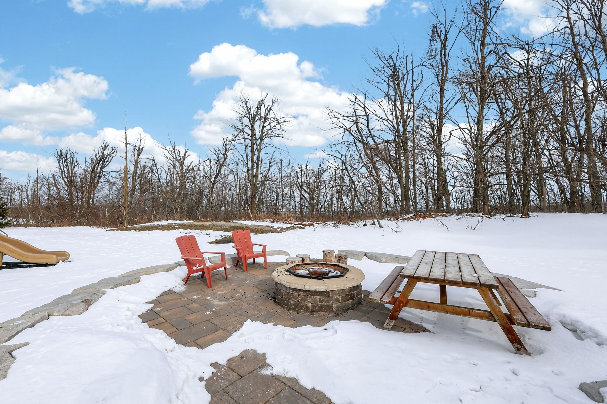 A snow-covered outdoor area with a fire pit, two red chairs, and a wooden picnic table.